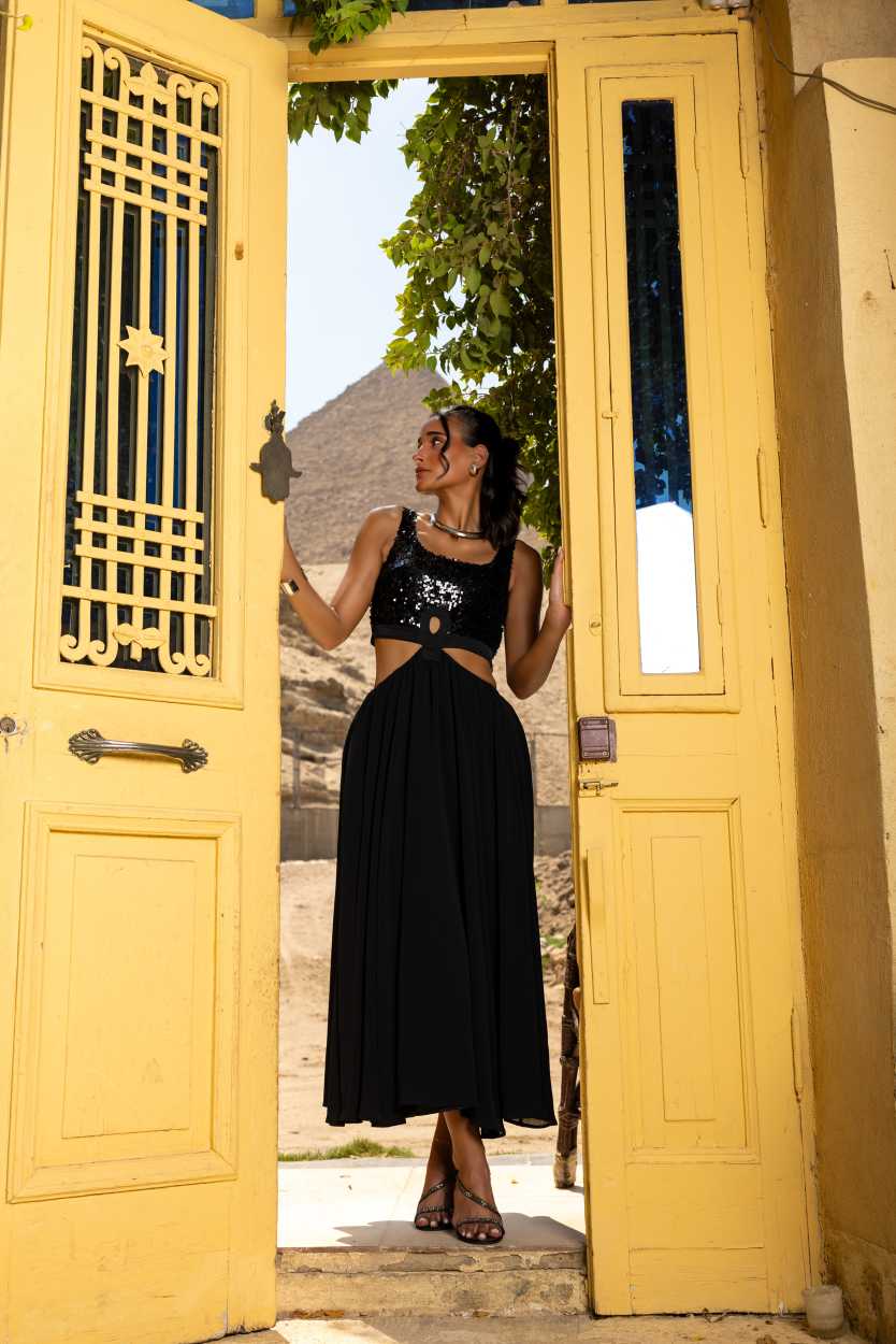 Woman in a black dress standing in an open yellow door with greenery outside Clep