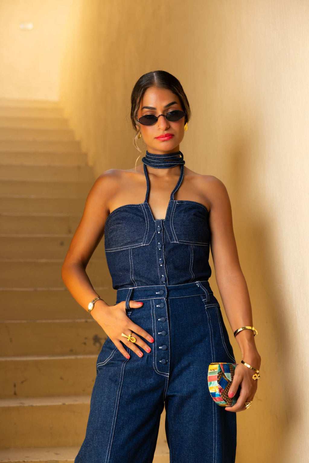 Woman wearing a denim jumpsuit with sunglasses and a clutch against a beige wall. Clep