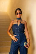 Woman wearing a denim jumpsuit with sunglasses and a clutch against a beige wall. Clep