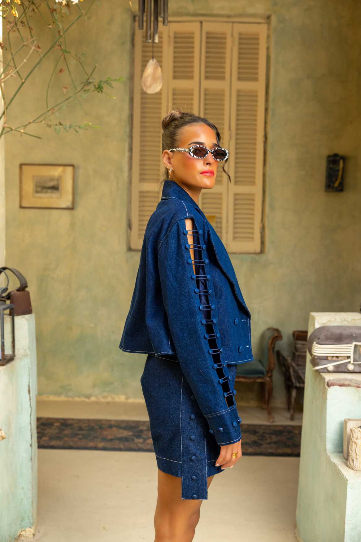 Woman wearing a blue denim outfit in an indoor setting with green walls and wooden shutters Clep