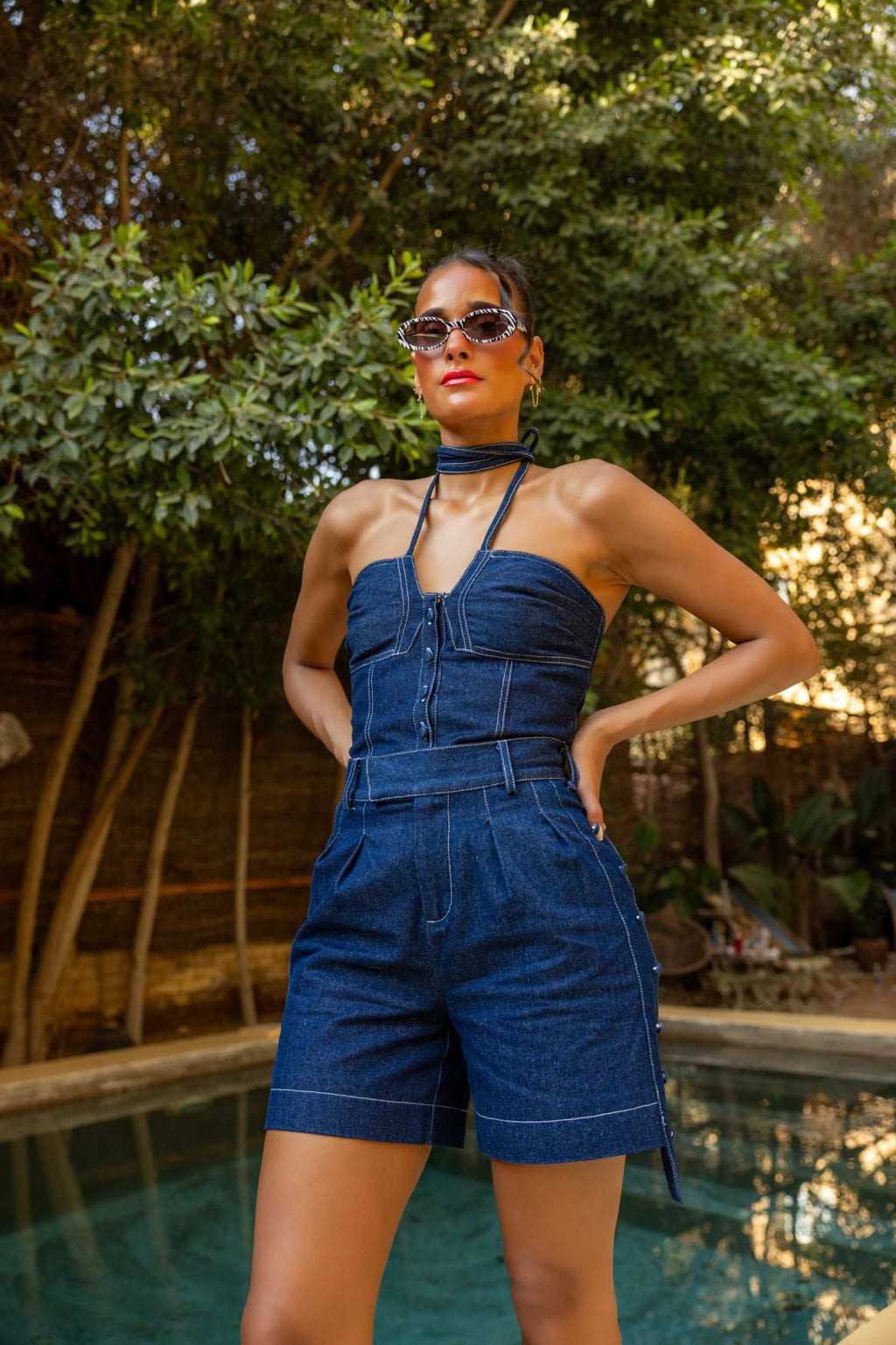 Woman in a denim jumpsuit standing by a pool with greenery in the background Clep