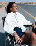 Woman sitting on a chair by a road, wearing a white shirt with a green design. Clep
