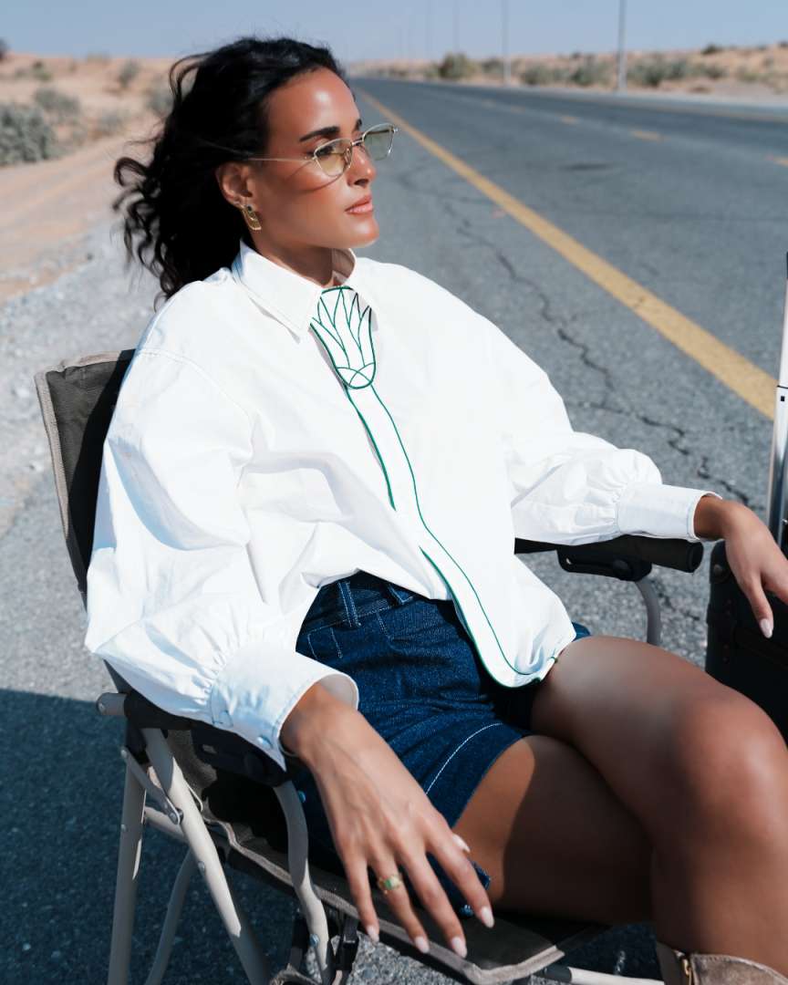 Woman sitting on a chair by a road, wearing a white shirt with a green design. Clep