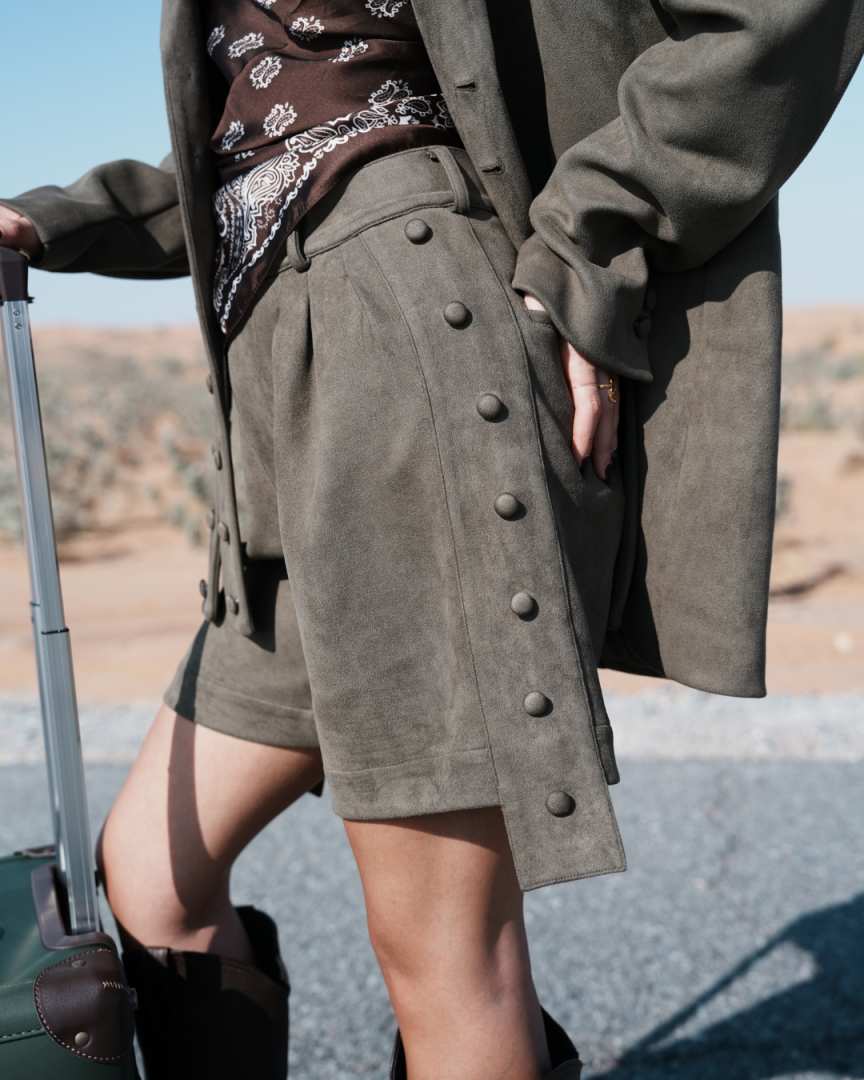 Person wearing a green coat and shorts with a suitcase in a desert setting Clep