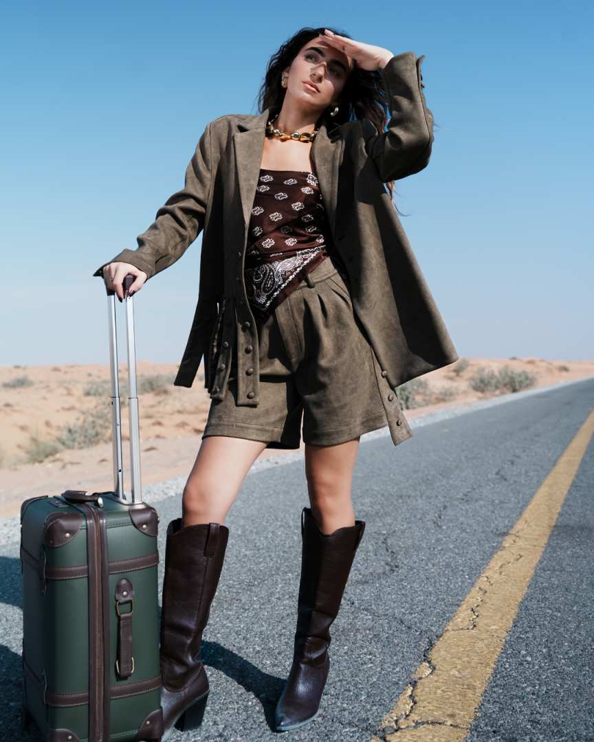 Woman in a desert setting with a suitcase, wearing a green coat, shorts, and boots. Clep