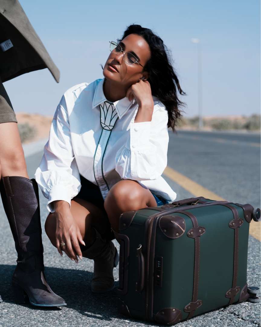 Woman in white shirt and boots sitting on a suitcase by a road Clep