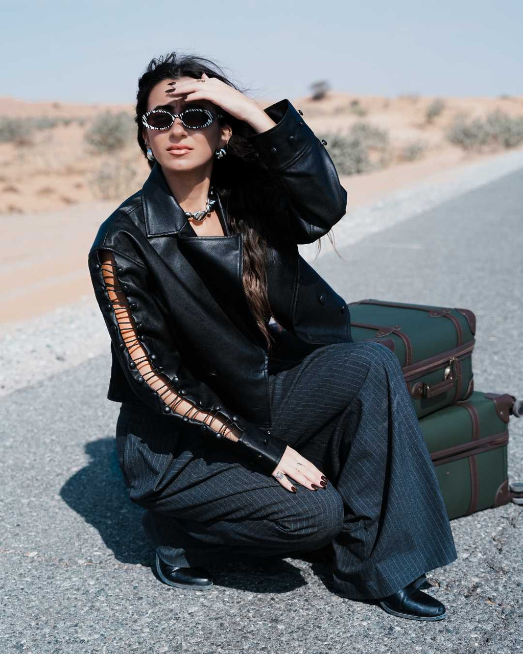 Woman in black outfit with leather jacket and sunglasses, sitting on a road with vintage suitcases. Clep