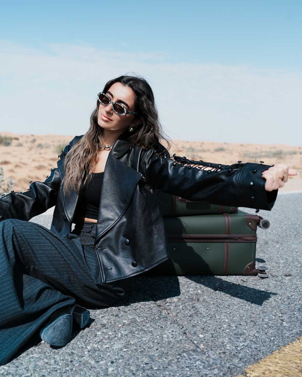 Woman in a black leather jacket sitting on the ground with a suitcase in the desert. Clep