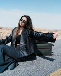 Woman in a black leather jacket sitting on the ground with a suitcase in the desert. Clep