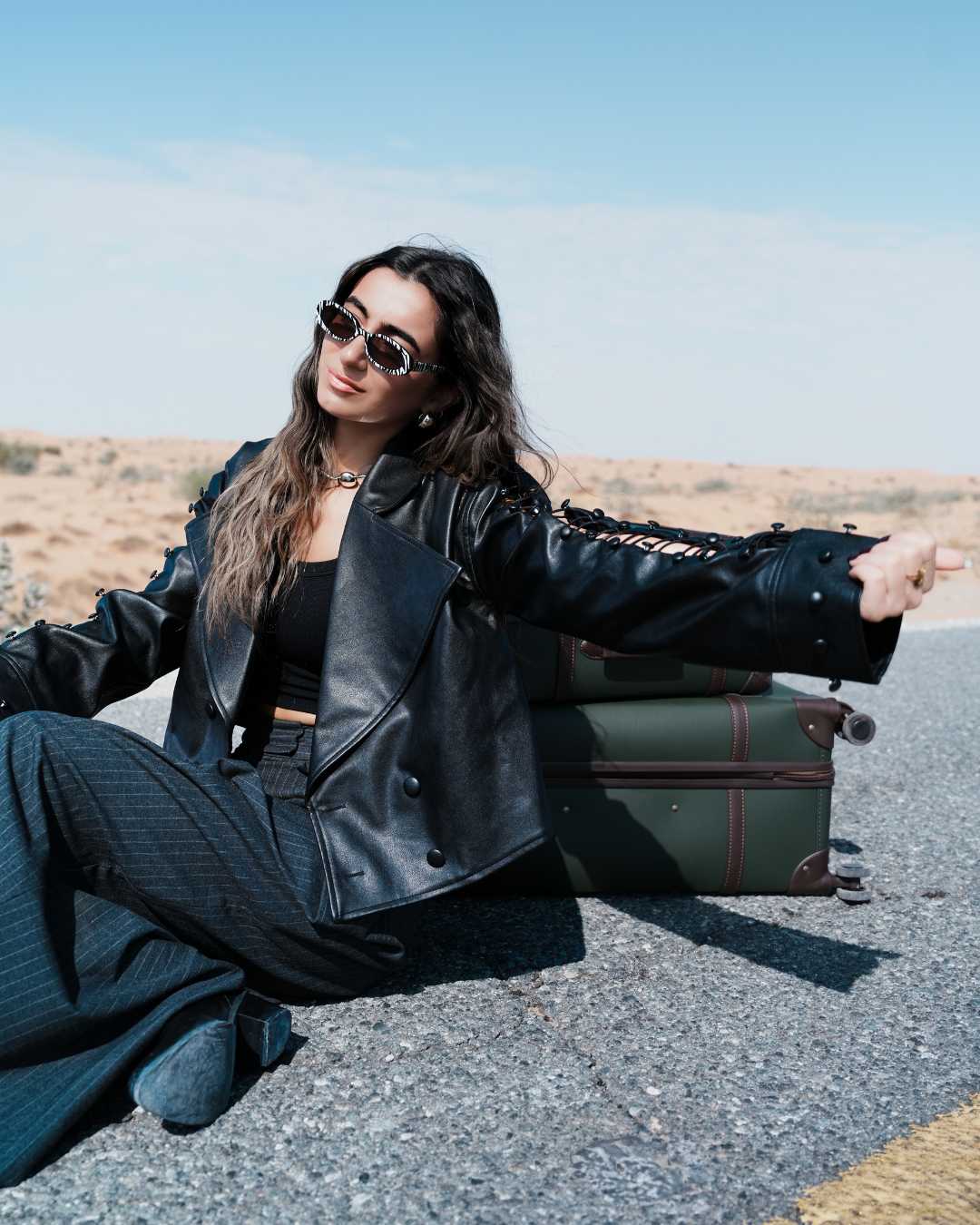 Woman in a black leather jacket sitting on the ground with a suitcase in the desert. Clep