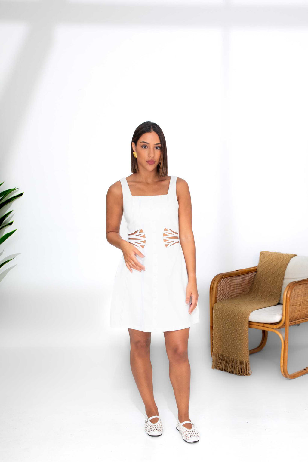 Woman wearing a white dress with brown patterns standing in a minimalistic room with a chair and plant. Clep