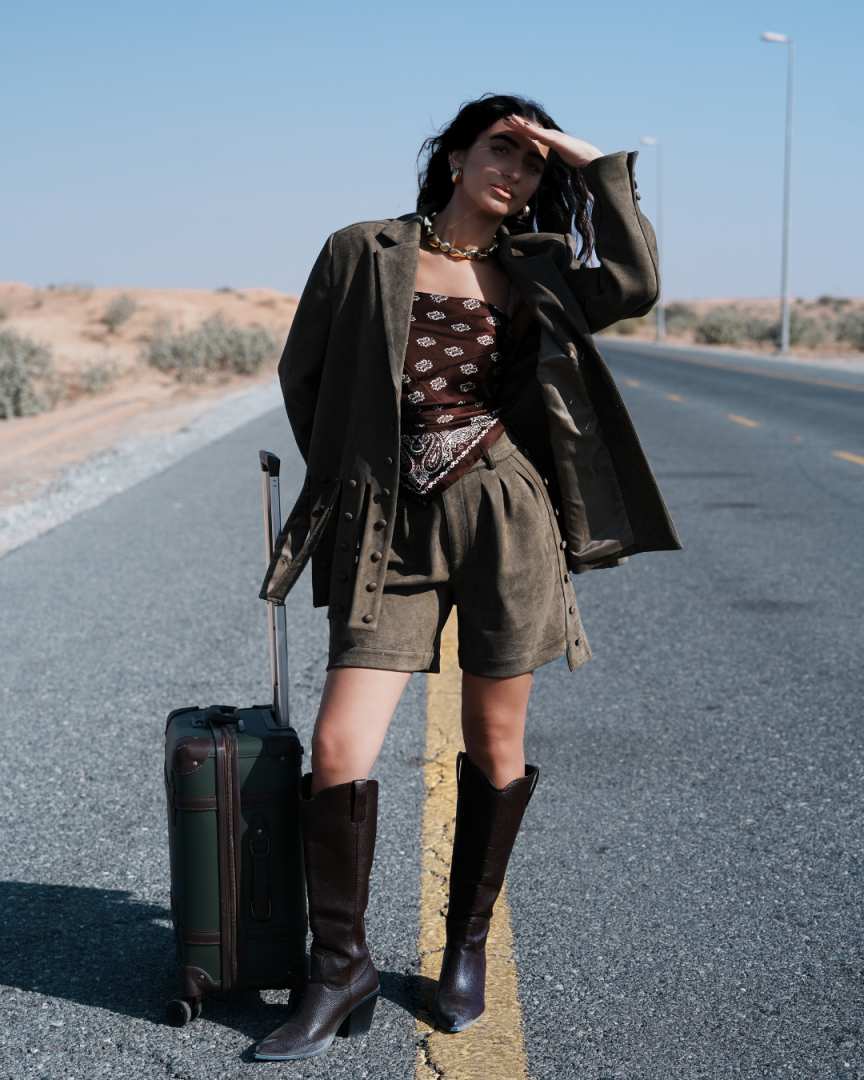 Woman in vintage-style outfit with leather jacket, boots, and suitcase on a desert road. Clep
