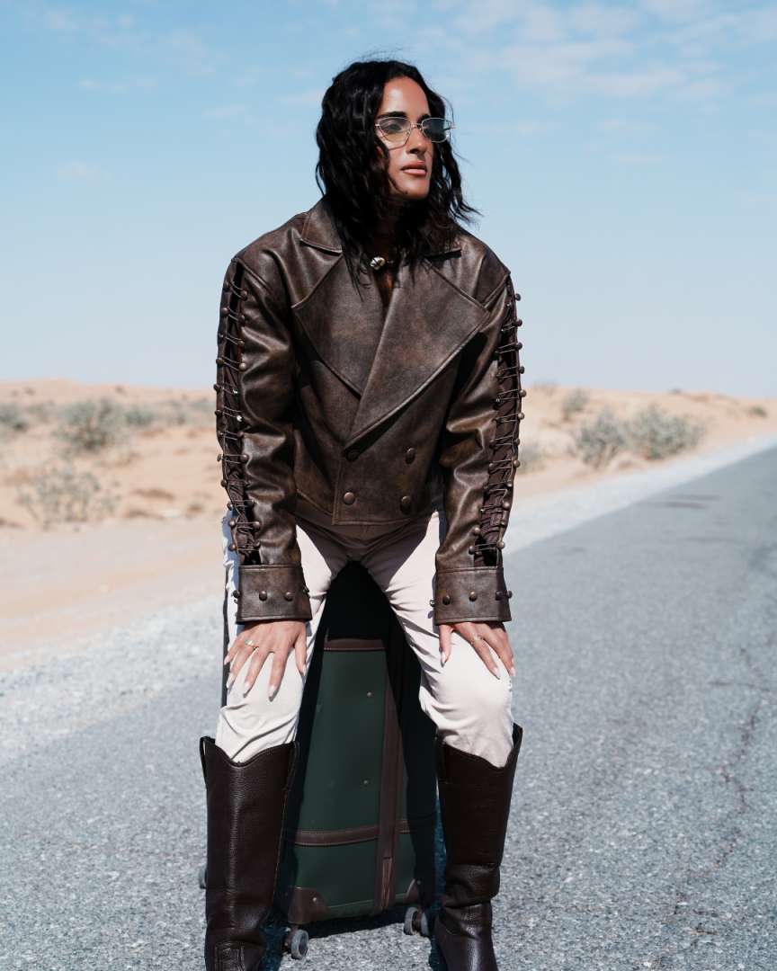Person wearing a brown leather jacket and white pants standing on a road with a suitcase. Clep