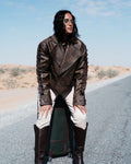Person wearing a brown leather jacket and white pants standing on a road with a suitcase. Clep