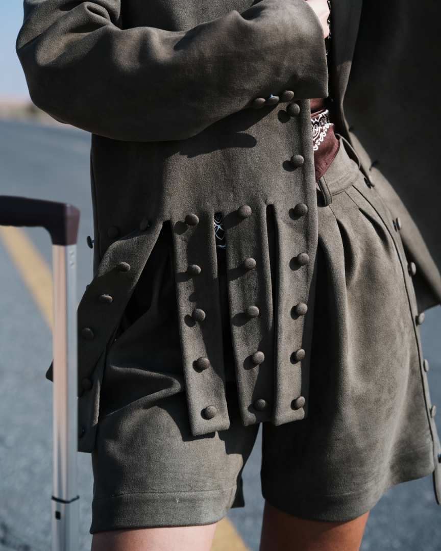 Person wearing a dark green coat with textured sleeves and a matching skirt, holding a suitcase. Clep