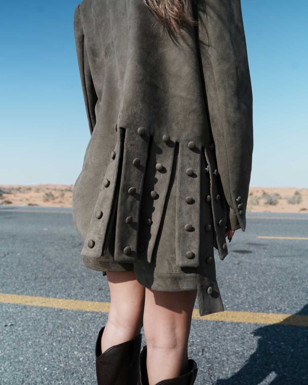 Person wearing a dark coat with button details on a road with desert landscape in the background Clep