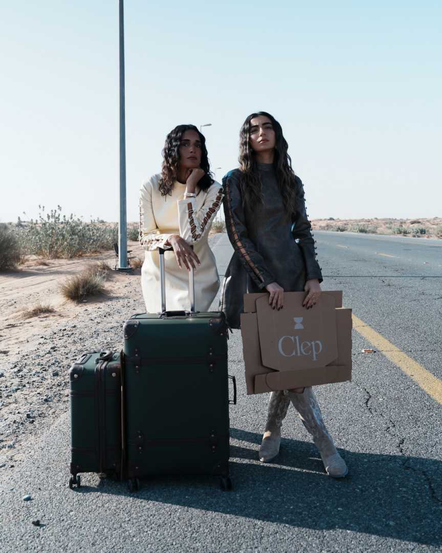 Two women standing on a desert road with luggage and a box labeled 'Clep'.
