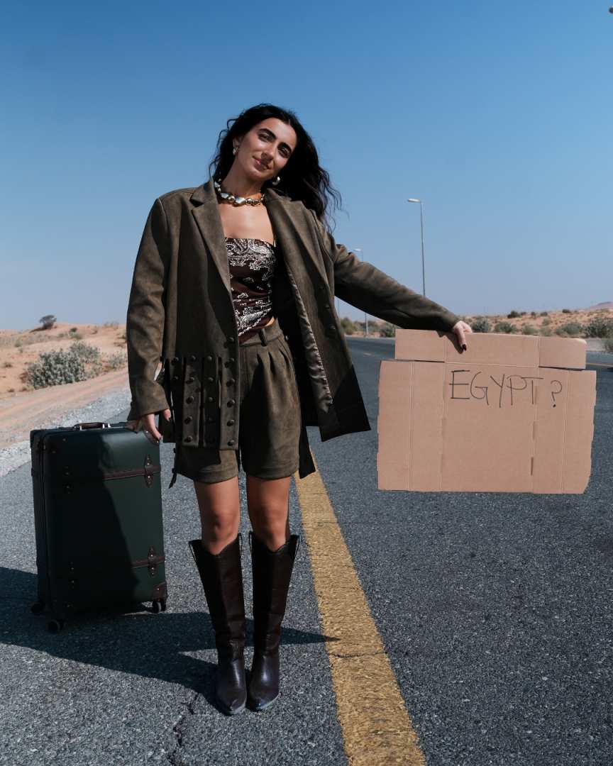Woman standing on a road with a suitcase and a cardboard sign asking 'Egypt?' Clep